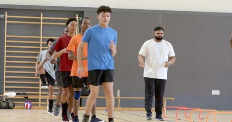 Diverse male athletes running drill in indoor gym while coach observing hurdle training