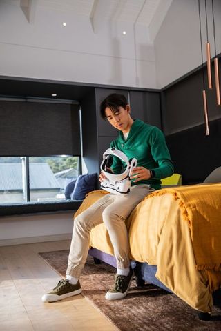 Asian man sitting on bed edge inspecting white astronaut helmet in modern bedroom daylight