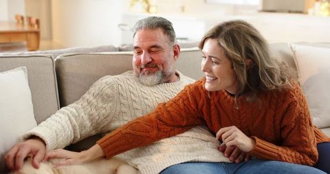 Middle-aged couple petting golden retriever on cozy beige sofa wearing cable-knit sweaters, smiling