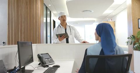 Asian doctor consulting receptionist in hijab at modern clinic desk with tablet
