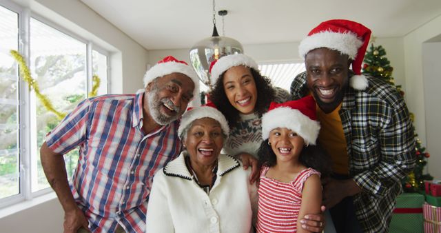 Multigenerational African American family celebrating Christmas at home. Everyone wears red Santa hats and smiles cheerfully in front of a decorated Christmas tree. Perfect for holiday-themed promotions, family celebration advertisements, and festive greeting cards.