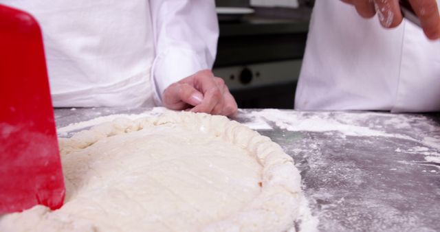 Chefs wearing white coats preparing pizza dough with red scraper in a professional kitchen. Ideal for content about culinary arts, food preparation, professional kitchens, team cooking efforts, and baking.