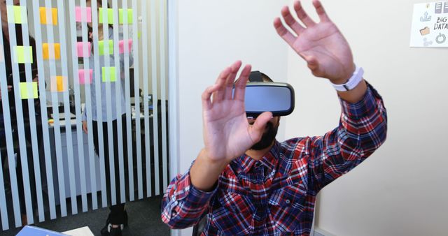 Man wearing VR headset in office workspace, arms raised, looking immersed in a virtual reality experience. In background, visible glass wall with sticky notes. Ideal for illustrating modern office settings, technology use in businesses, innovation in workplace, and creative working environment.