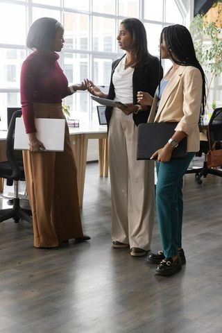 Diverse women collaborating in modern open-plan office holding laptop, tablet and folio