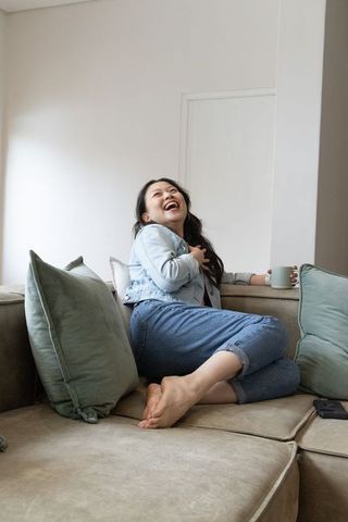 Joyful asian woman laughing on cozy beige sofa holding mug in minimalist living room