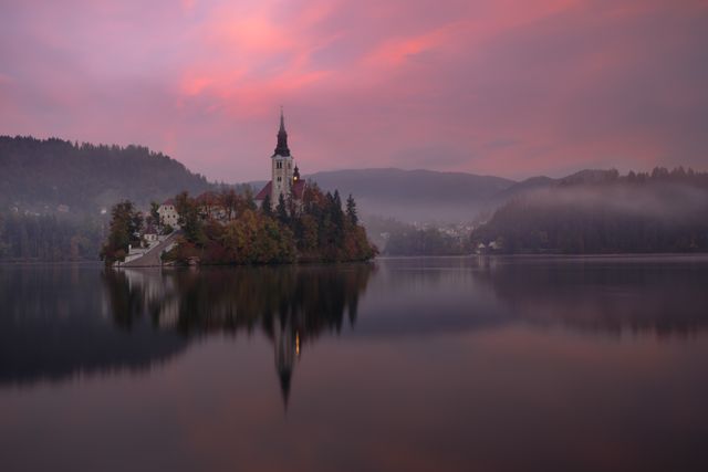 Church on Lake Bled at sunrise with a vibrant pink sky. Calm water perfectly mirrors the island and surrounding landscape, creating a peaceful scene. Mist adds to the ethereal atmosphere. Ideal for travel articles, nature magazines, and contemplative spaces.