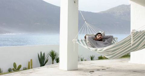 Indian man reclining in striped hammock on seaside veranda relaxing overlooking ocean