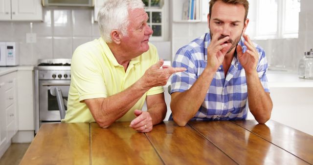 Senior father engaging in a serious discussion with his adult son in a modern kitchen. Both are seated at a wooden table, emphasizing familial bonds and communication. Ideal for use in content relating to family relationships, parental advice, communication strategies, or mentoring dynamics.