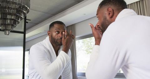 Mature black man applying eye cream at mirror wearing white bathrobe in modern bathroom