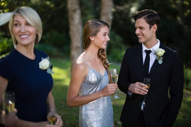 Couple celebrating their wedding with champagne in a park. The bride is wearing a silver dress and the groom is in a black suit. They are smiling and enjoying the moment with a guest. Perfect for use in wedding planning materials, celebration announcements, and event promotions.