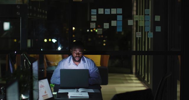 Man working late at night in modern office, focused on laptop with documents and sticky notes on window. Perfect for use in articles about late-night work culture, deadline pressures, productivity, and corporate environments.