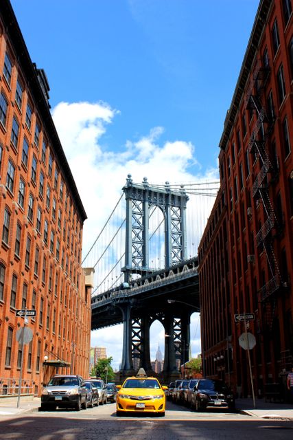 View of Manhattan Bridge framed by historic brick buildings and yellow taxi on cobblestone street in New York. Perfect for illustrating urban life, travel to New York, and showcasing famous landmarks. Suitable for travel blogs, city guides, tourism promotional material, posters, and postcards.