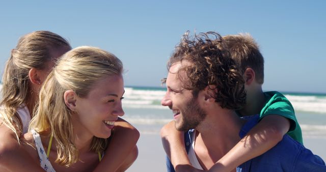 A happy family enjoying their summer vacation, having fun with a piggyback ride on a sandy beach with the ocean in the background. Ideal for advertisements or promotional materials for travel agencies, family retreats, summer activities, or beach resort marketing campaigns.