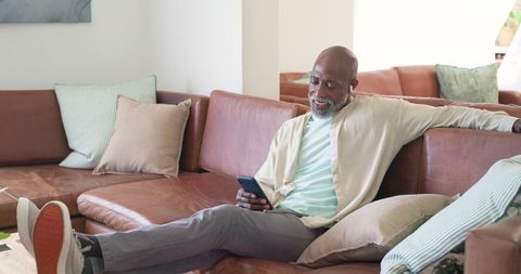 Senior african american man relaxing on leather sofa at home using smartphone and smiling