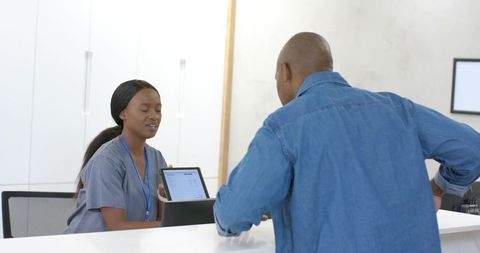 African female nurse assisting patient at clinic reception using tablet for check-in