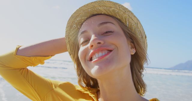 A young Caucasian woman smiles brightly while enjoying the sun at the beach, with copy space. Her straw hat and radiant expression convey a sense of joy and relaxation.