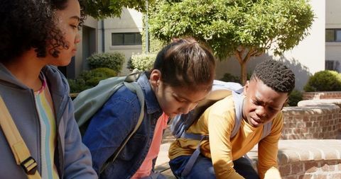 Concerned diverse teenagers sitting on brick courtyard ledge with backpacks, school campus