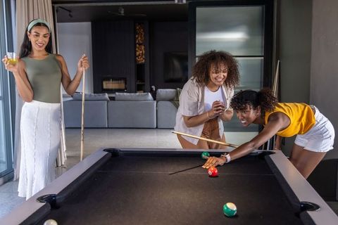 African american women playing pool in modern living room laughing during game night