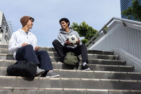 Diverse teen friends sitting on urban stairs holding soccer ball and backpacks