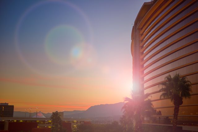 Beautiful sunset scene with high-rise hotel building and palm trees in foreground, capturing vibrant sunlight and sun flare, with distant mountains and clear sky. Ideal for travel advertisements, urban landscape illustrations, tourism marketing, hotel and hospitality promotions.