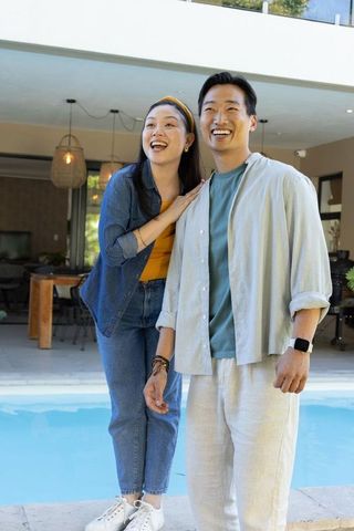 Smiling asian couple enjoying poolside patio at modern home, casual summer lifestyle