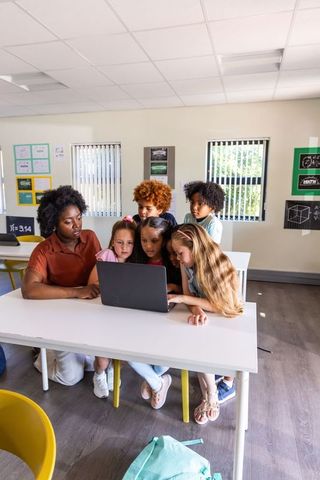 Female Teacher and Diverse Elementary Students Collaborating on Laptop in Bright Classroom