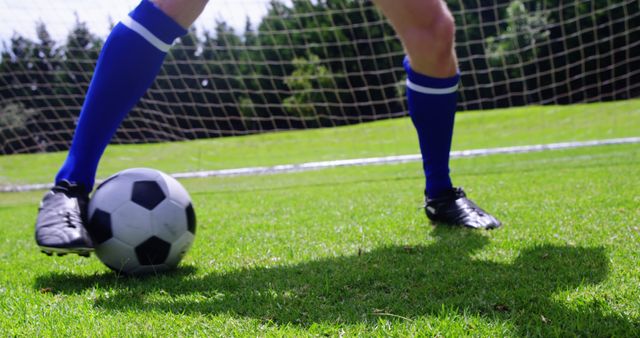 A soccer player in black cleats is about to kick a soccer ball on a lush green field, with copy space. Capturing the action and energy of a soccer game, the focus on the player's feet and ball emphasizes the skill and precision involved in the sport.