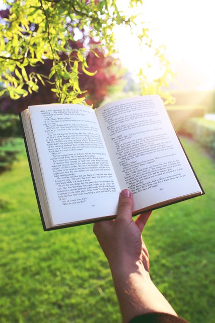 This image shows a person holding an open book outdoors with bright sunlight streaming through foliage. Suitable for content related to reading, education, relaxation in nature, and promoting literacy. Ideal for use in blogs, articles, websites about self-improvement, education, and outdoor activities.