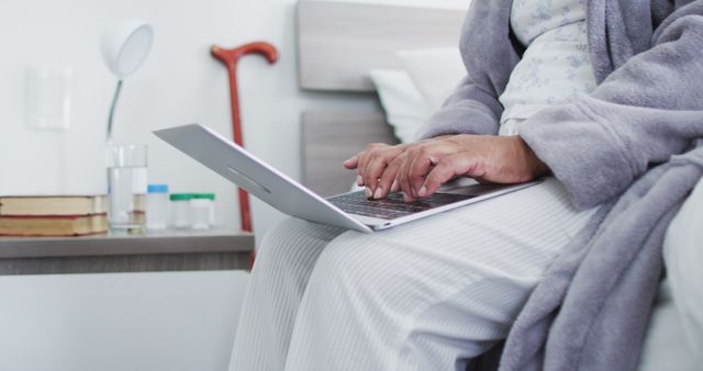 Elderly woman using laptop while sitting on bed, with focus on hands typing. This image portrays the use of technology among seniors, highlighting their engagement in modern communication and digital activities. Ideal for illustrating concepts related to senior lifestyle, independence, remote communication, technology adoption by elderly, or promoting assistive technology services.
