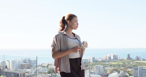 Young woman enjoying tea on coastal balcony, gazing at city skyline and sea