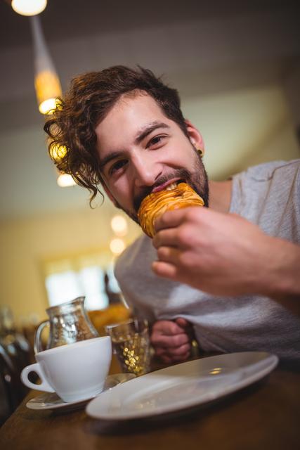 Young man relishing a croissant while seated in a cozy café. Perfect for marketing food and beverage establishments, lifestyle blogs, or social media promotions focusing on casual and enjoyable dining experiences.