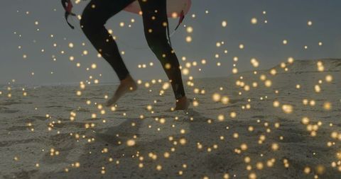 Surfer walking on sandy beach in wetsuit carrying surfboard with golden bokeh lights