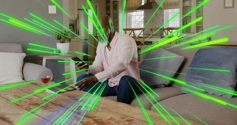 Man on living room sofa holding phone reaching for laptop with neon green light bursts
