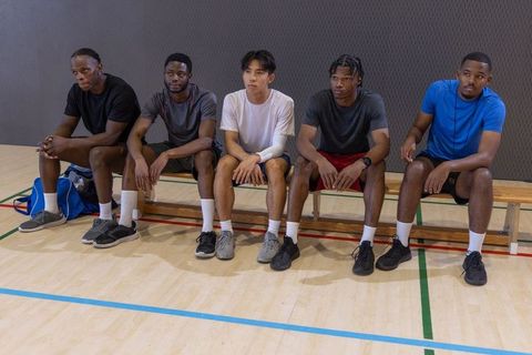 Diverse male athletes sitting on bench in gym court waiting between basketball drills