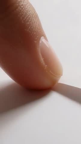 Vertical macro video of fingertip sliding along paper edge with glossy nail and shadow