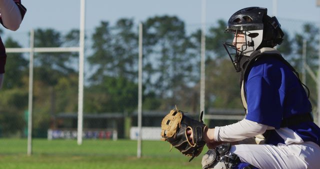 A baseball catcher in full gear crouching and waiting for the pitch during an outdoor game on a sunny day. Suitable for use in marketing materials related to sports equipment, baseball events, athletic coaching, and youth sports promotion. Ideal visual for articles discussing baseball techniques, training, and team sports for youth and adults.