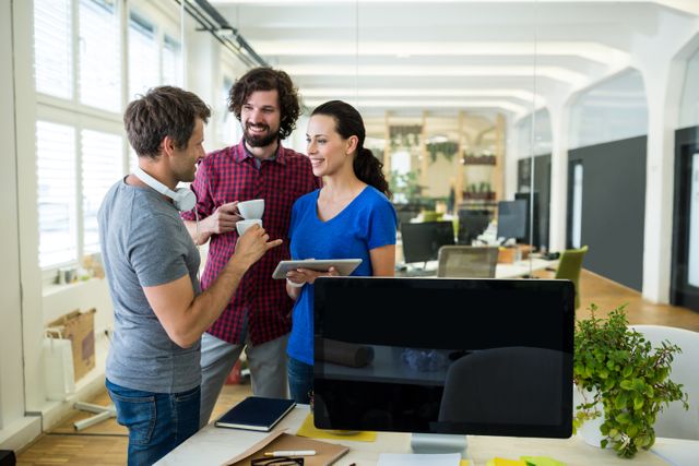 Three colleagues are engaging in a collaborative discussion in a modern, bright office environment. They are dressed casually, indicating a relaxed work atmosphere conducive to creativity. One individual holds a digital tablet, suggesting they are reviewing a project or brainstorming ideas. This image can be used to depict teamwork, modern office culture, creative industries, and collaborative workspaces in business and marketing materials.