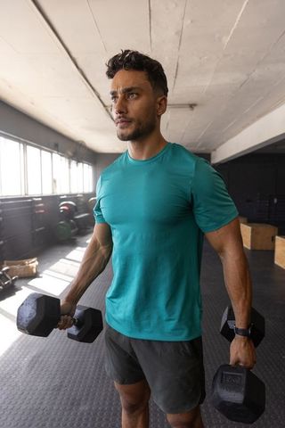 Middle eastern man lifting hex dumbbells in industrial gym wearing teal shirt workout