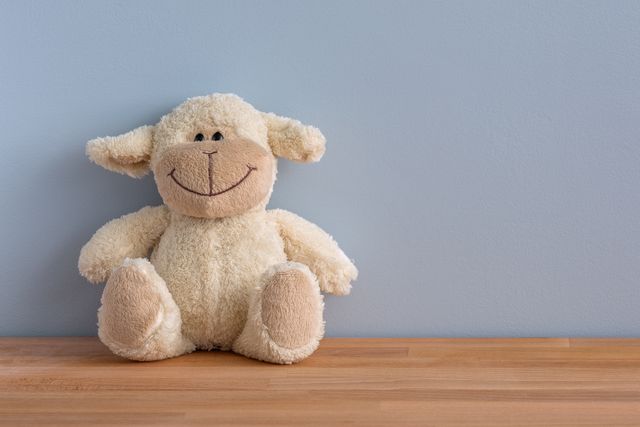 Smiling sheep stuffed toy with fluffy fur and large embroidered smile sitting on wooden table against light blue wall. Ideal for adding a touch of warmth and charm to nurseries, children's bedrooms, or play areas. Great for use in advertisements for baby products, gift shops, or as a cute addition to promotional material for toy stores and parenting blogs.