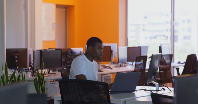 Young man in casual attire working on a computer at a modern office with an orange accent wall. Ideal for themes related to business, productivity, technology, and modern work environments. Perfect for use in articles, presentations, marketing materials, or any content that illustrates a contemporary office setup and workspace.