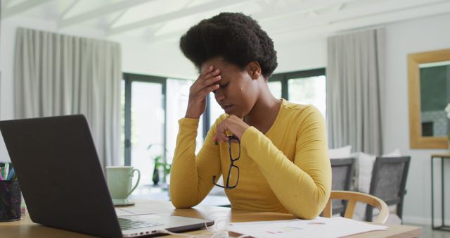 Woman experiencing stress while working from home, sitting at desk with laptop and holding glasses. May be used to depict work-related stress, remote work challenges, telecommuting issues, or general office frustrations.