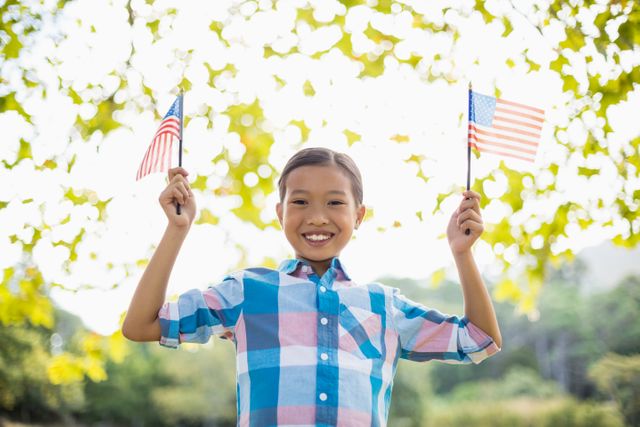 Young girl holding American flags in both hands, smiling brightly in a park. Ideal for use in content related to patriotism, national holidays like Independence Day, family gatherings, outdoor activities, and children's events. Perfect for promoting community events, educational materials, and festive celebrations.