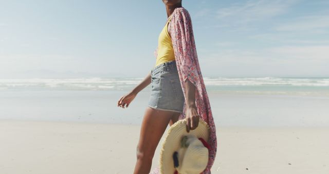 Woman in a yellow top, denim shorts, and a floral kimono walking along sandy beach holding a straw hat. Ideal for promoting summer fashion trends, vacation destinations, apparel brands, or lifestyle blogs focusing on relaxation and beach activities.