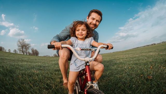 Father guiding young daughter riding bike in sunlit meadow smiling and laughing together