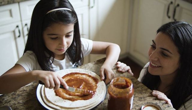 Mom teaching daughter spreading tomato sauce on pizza crust in bright home kitchen