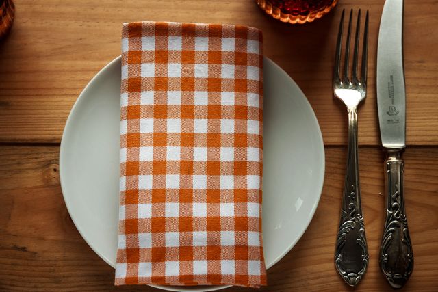 Ideal for designs related to dining and hospitality themes, this scene showcases a well-arranged rustic table setting. It features a checkered napkin neatly folded over a white plate, placing emphasis on the elegance of table decor. The ornate knife and fork add a classic, vintage touch, while the wooden table creates a warm, inviting atmosphere perfect for advertisements for restaurants, blogs about home decor and culinary arts, or cookbooks. Great for illustrating meal preparation and refined dining experiences.