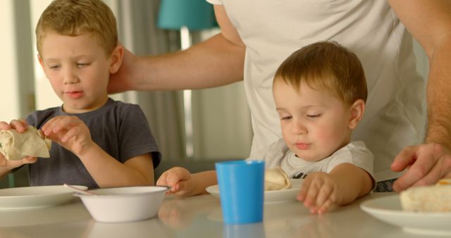 Father bonding with his young sons at home during lunchtime. The boys eat a meal together, showcasing family love and togetherness. Ideal for use in articles about parenting, familial relationships, healthy eating at home, or home life. Promotes themes of family time, parent-child interaction, and domestic routines.