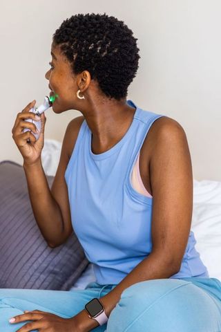African american woman using handheld led oral-care device on bed wearing smartwatch