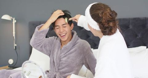 Smiling couple relaxing in bathrobes on bed while woman rolling jade facial roller on man