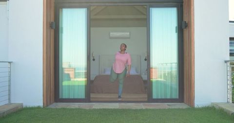African american woman stretching at open glass bedroom doorway overlooking terrace lawn